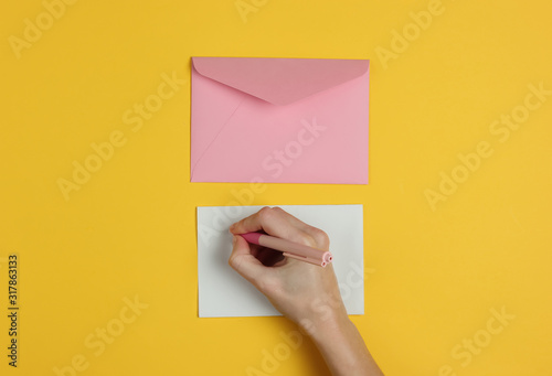 Female hand writes a letter with a pen on yellow background. Wedding invitations, love letter, valentine. Top view