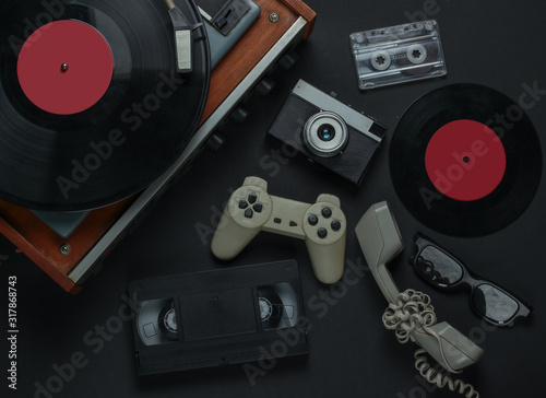 Flat lay retro media and entertainment. Vinyl record player with vinyl record, film camera, video cassette, audio cassette, gamepad, handset on a black background. 80s. Top view