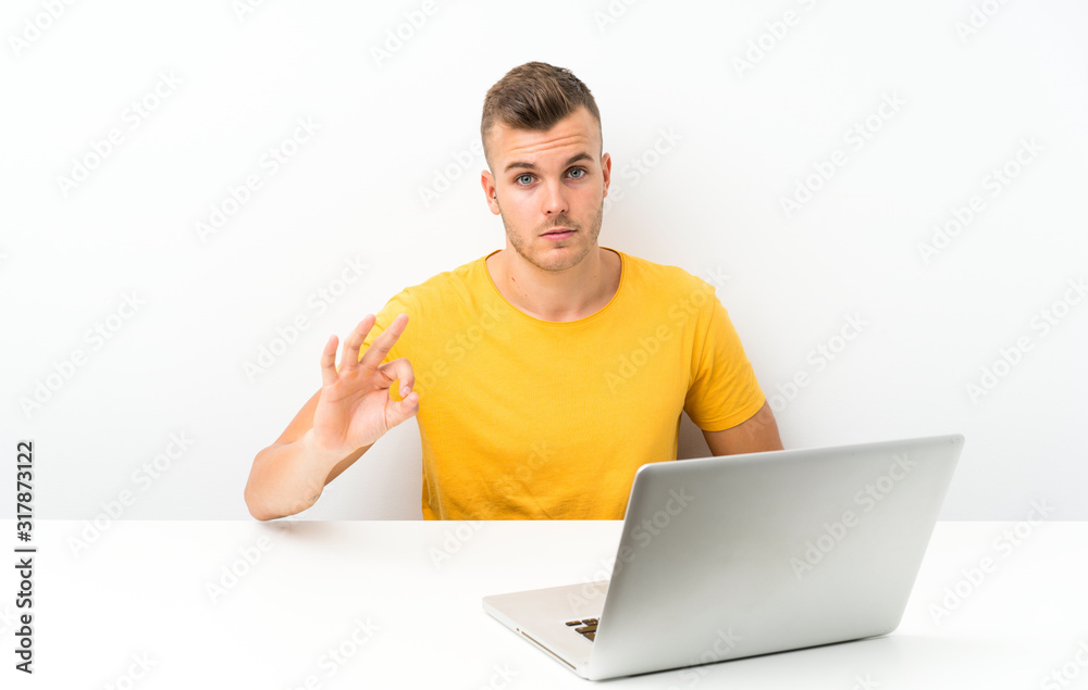 Young blonde man in a table with a laptop showing an ok sign with fingers Young blonde man in a table with a laptop showing an ok sign with fingers