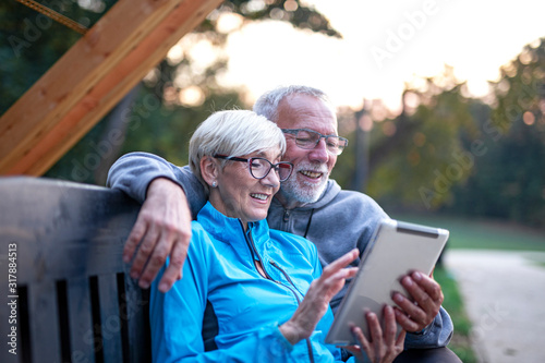 Mature couple sitting at banch in par and looking at tablet and having fun