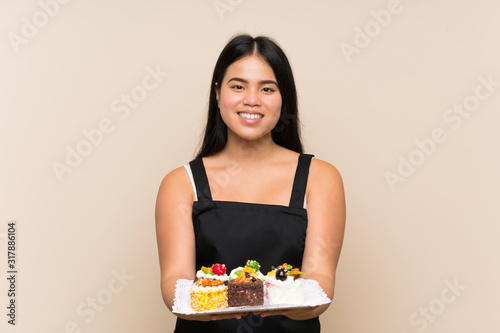 Young teenager Asian girl holding lots of different mini cakes over isolated background