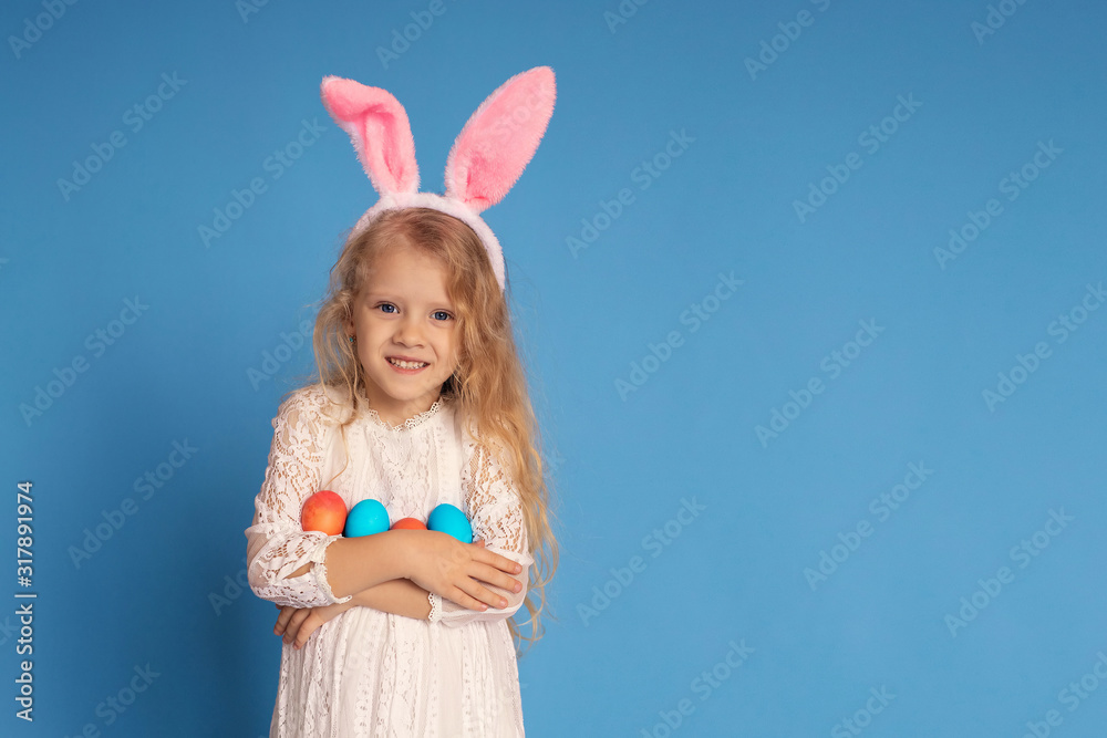 Portrait of a cheerful laughing girl in a white dress and pink rabbit ears holding Easter eggs in her hands. isolated on a blue background