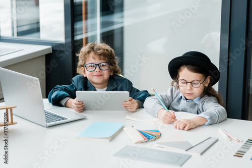 Cute schoolboy with tablet looking at you while sitting next to classmate
