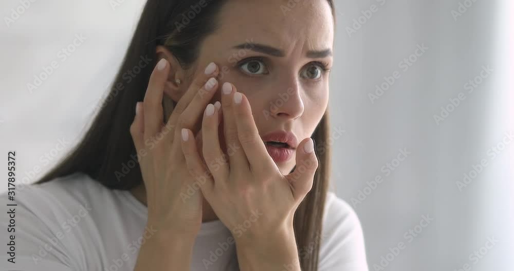 Worried woman looking in mirror, examining wrinkles in eyes area.