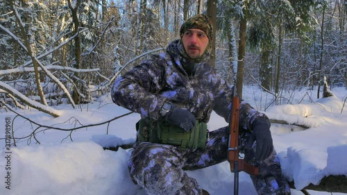 A frosty day in a winter snowy forest. The hunter in a camouflage suit sat down to rest on a fallen tree and drink hot tea.