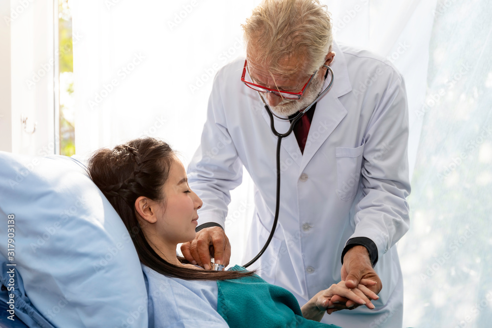 Senior professional Caucasian man examining Asian woman patient with stethoscope in the hospital room.