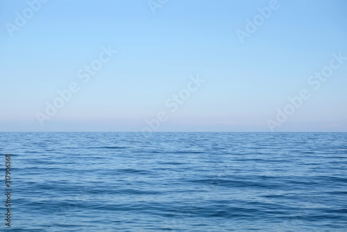 Calm sea surface with small waves in the morning and the land on skyline in light haze in pink and blue colors under clear cloudless sky horizontal view © DyMaxFoto