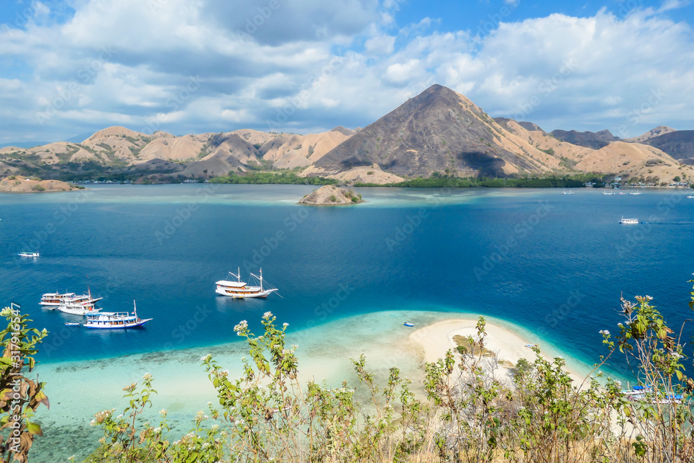 Naklejka premium View from top of a cliff on Kelor Island, Komodo, Indonesia. Island is surrounded with white sand beaches and turquoise water. There is another island in the back.There are boats anchored to a shore.
