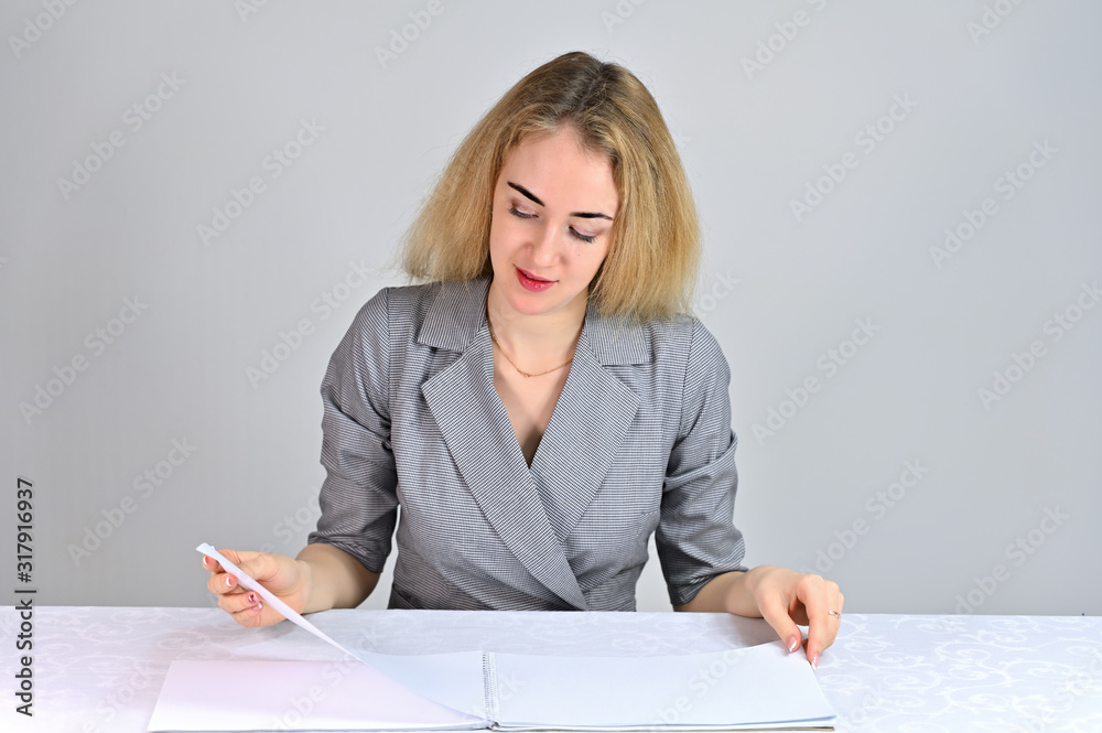 Model Sits at a table directly opposite the camera in various poses. Portrait of a pretty cute smiling young blonde business woman with minimal makeup in a gray suit on a white background. Model Sits at a table directly opposite the camera in various poses. Portrait of a pretty cute smiling young blonde business woman with minimal makeup in a gray suit on a white background.