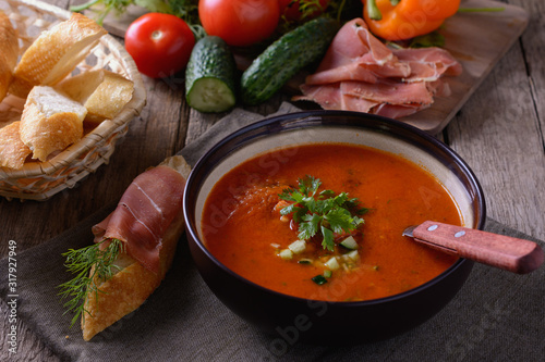 Tasty gazpacho tomato soup in a plate and jamon on a slice of baguette on a dark napkin on a wooden table next to vegetables and slices of bread