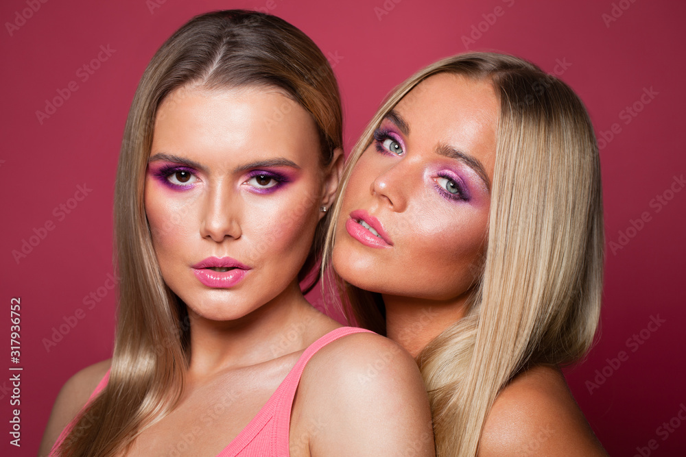 Two blonde women fashion models with long straight hair and makeup on pink background