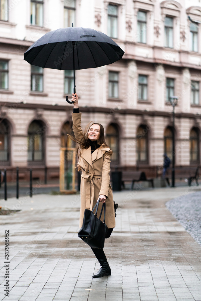 Outdoor photo of brunette lady jumping with black umbrella in rainy autumn day.Fashion street style portrait. wearing dark casual trousers, white sweater and creamy coat.Fashion concept.