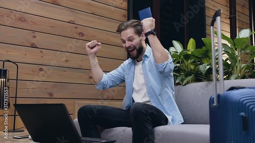 Handsome joyful bearded man sitting on couch in lobby looking on computer screen and waiting for result and after raised happily his hands up photo