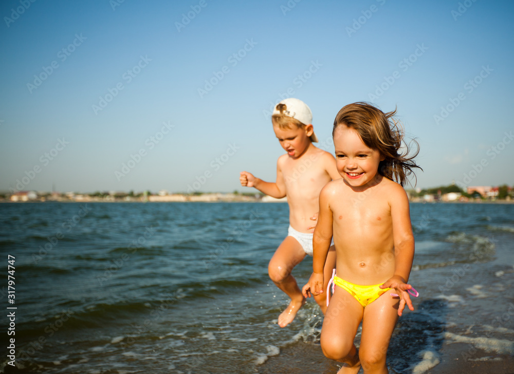 Happy little girl in bright swimwear and boy running on wavy sea water edge in sunshine and smiling on summer day. Family vacations and travelling concept Happy little girl in bright swimwear and boy running on wavy sea water edge in sunshine and smiling on summer day. Family vacations and travelling concept
