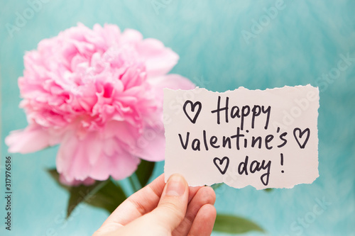 Happy Valentine’s Day - hand with card with lettering and pink peony flower on blue background