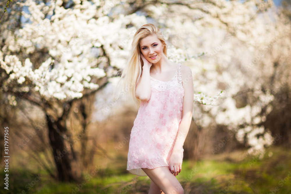 Young beautiful blonde smiling woman in white mini dress standing with blooming trees at background on clear sunny day. Woman natural beauty and summer nature concept Young beautiful blonde smiling woman in white mini dress standing with blooming trees at background on clear sunny day. Woman natural beauty and summer nature concept