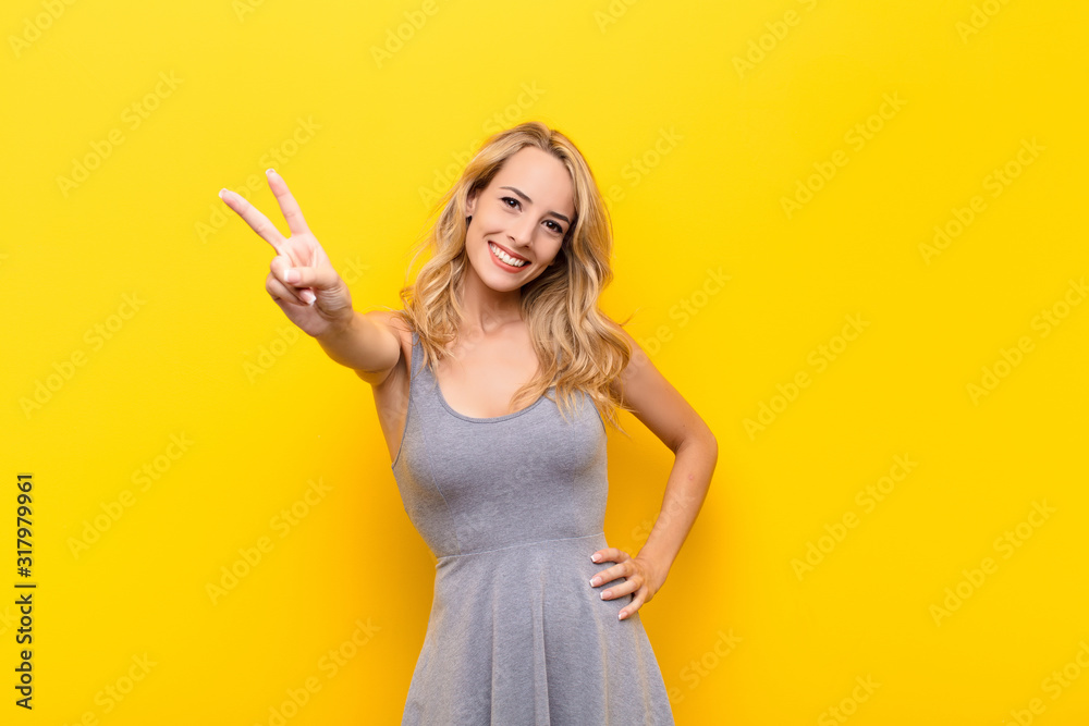 young blonde woman smiling and looking happy, carefree and positive, gesturing victory or peace with one hand against orange wall - obrazy, fototapety, plakaty 