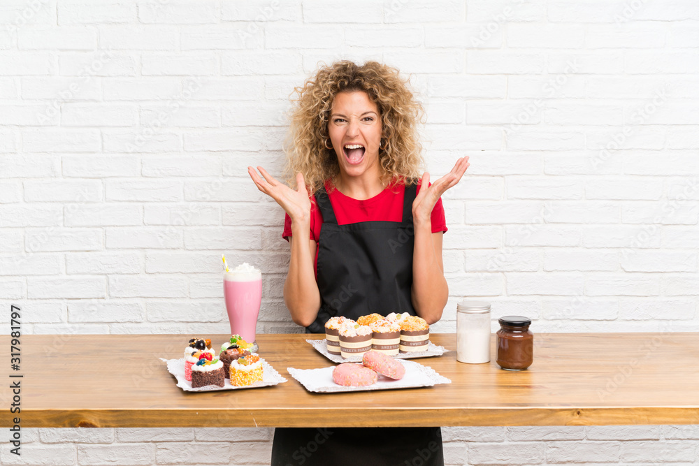 Young woman with lots of different mini cakes in a table unhappy and frustrated with something Young woman with lots of different mini cakes in a table unhappy and frustrated with something