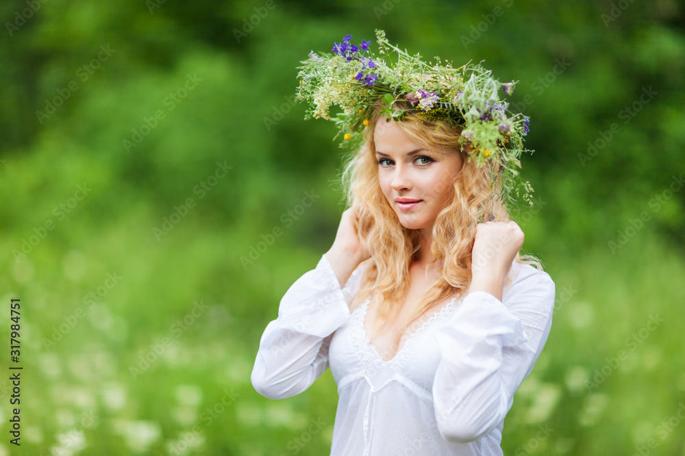Young beautiful blond woman in white dress and floral wreath standing and smiling on summer day Young beautiful blond woman in white dress and floral wreath standing and smiling on summer day
