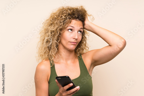 Young blonde woman with curly hair using mobile phone having doubts and with confuse face expression