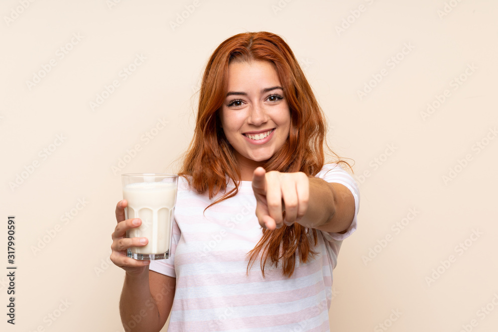 Teenager redhead girl holding a glass of milk over isolated background points finger at you with a confident expression