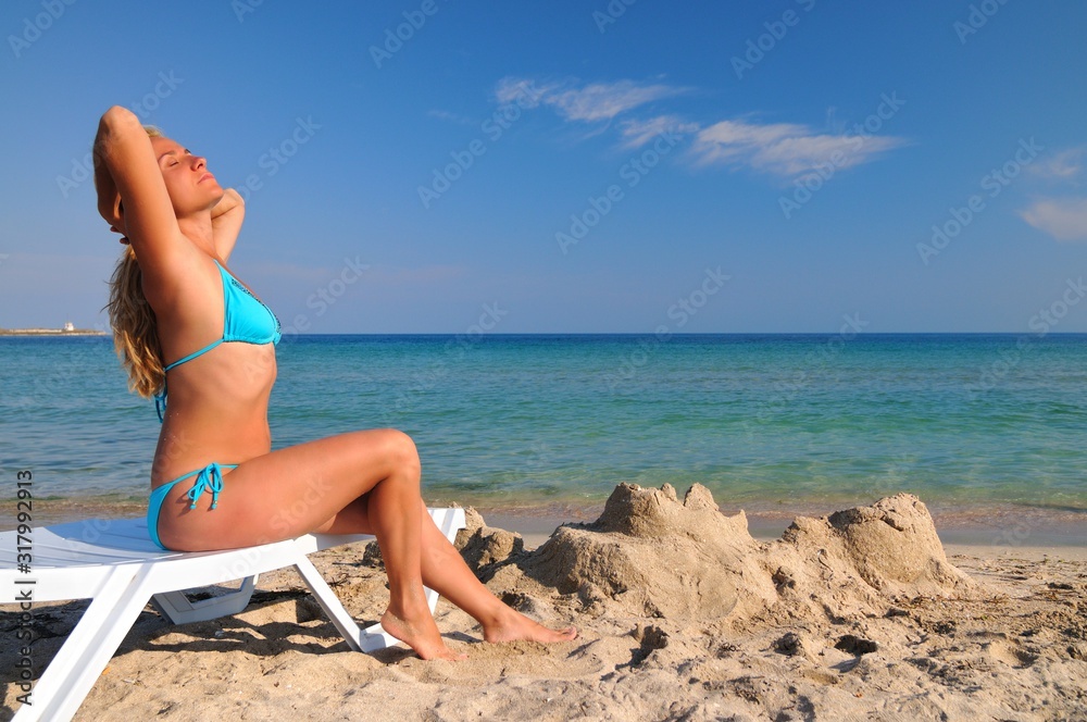 Young woman in bikini lying on sunbed near still sea water edge and enjoying sunshine Young woman in bikini lying on sunbed near still sea water edge and enjoying sunshine