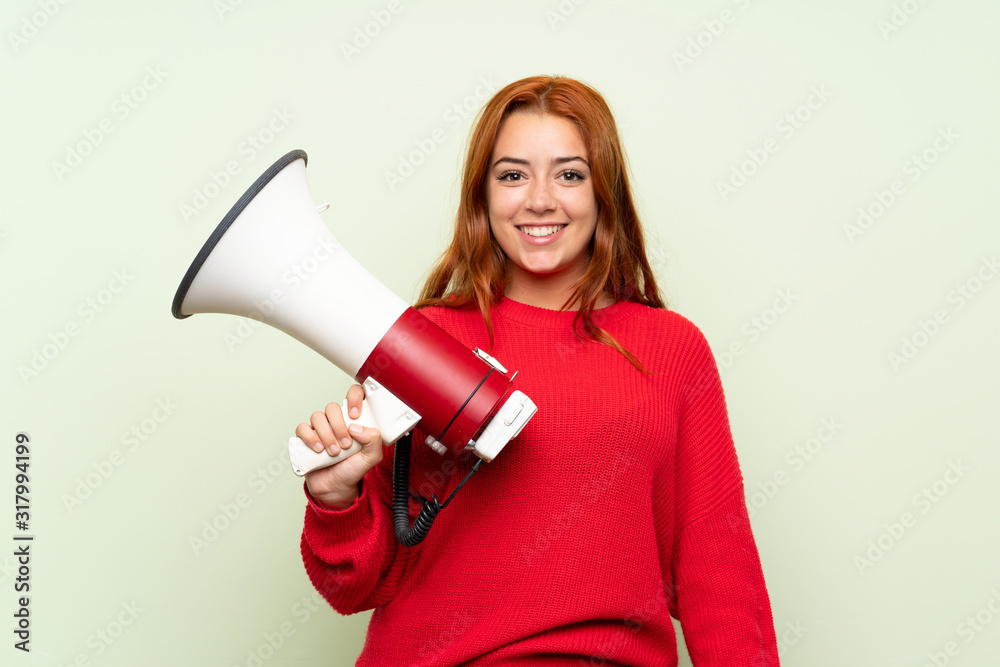 Teenager redhead girl with sweater over isolated green background holding a megaphone Teenager redhead girl with sweater over isolated green background holding a megaphone