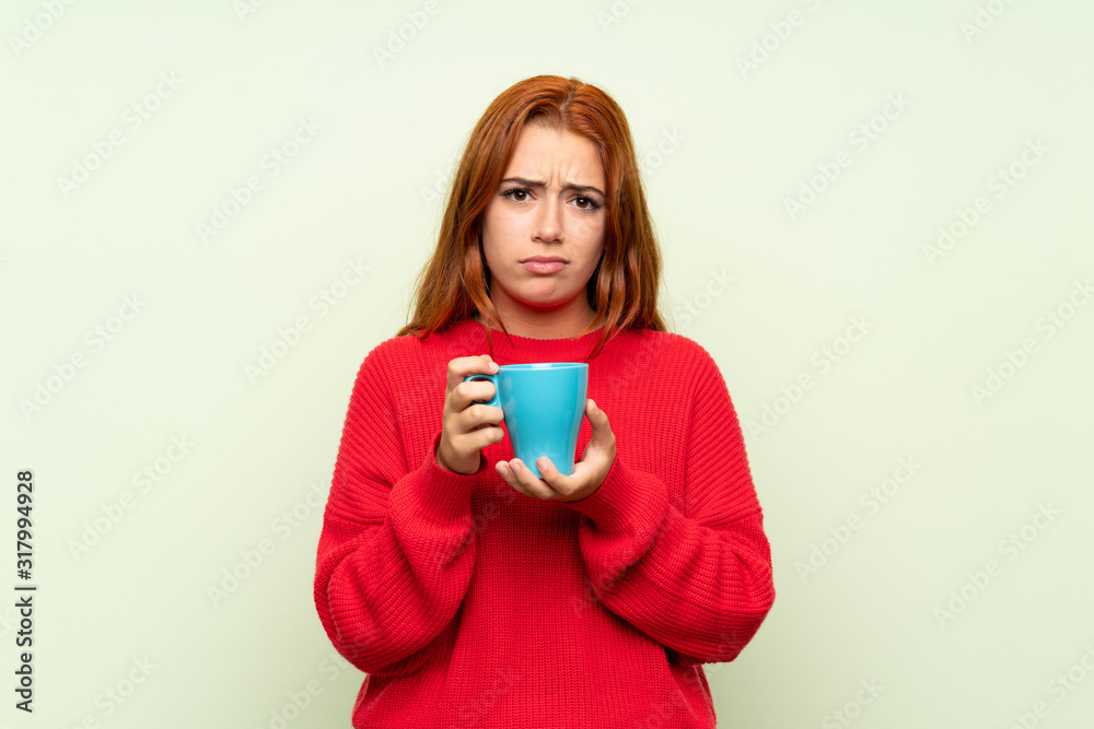 Teenager redhead girl with sweater over isolated green background holding hot cup of coffee Teenager redhead girl with sweater over isolated green background holding hot cup of coffee