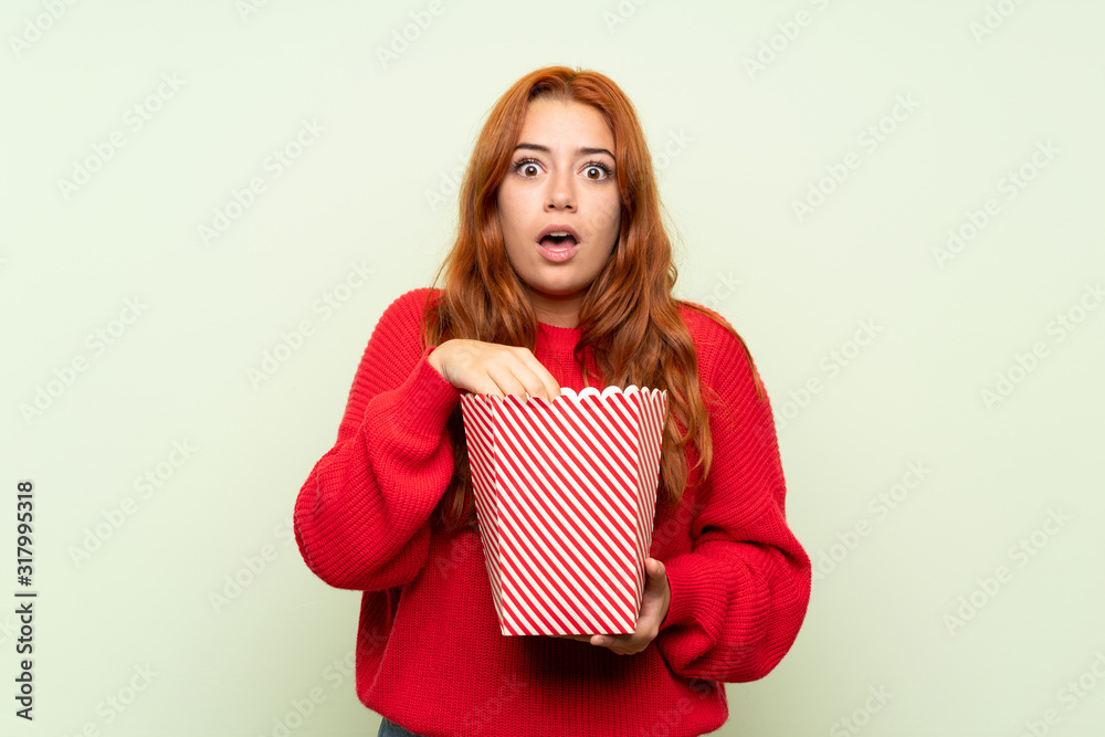 Teenager redhead girl with sweater over isolated green background eating popcorns Teenager redhead girl with sweater over isolated green background eating popcorns