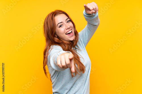 Teenager redhead girl over isolated yellow background points finger at you while smiling