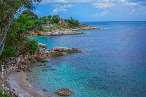 Beautiful landscape – sea coast: turquoise calm water, stones and rocks on the beach, cloudy sky and green trees. Corfu Island, Greece, Kassiopi village © elenakirey