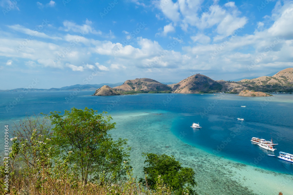 View from top of a cliff on Kelor Island, Komodo, Indonesia. Island is surrounded with white sand beaches and turquoise water. There is another island in the back.There are boats anchored to a shore.