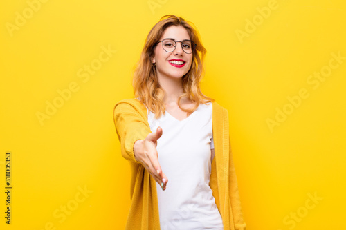 young pretty blonde woman smiling, looking happy, confident and friendly, offering a handshake to close a deal, cooperating against yellow wall