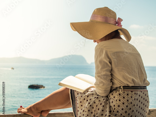 Young woman reading a book sitting on wooden balcony on Palmarola island in front of the ocean on a sunny day. Elegant white dress with skirt and hat. photo