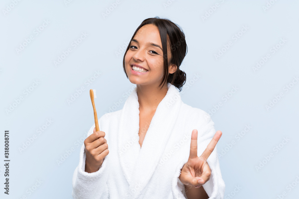 Young brunette woman in bathrobe brushing her teeth over isolated blue background smiling and showing victory sign Young brunette woman in bathrobe brushing her teeth over isolated blue background smiling and showing victory sign