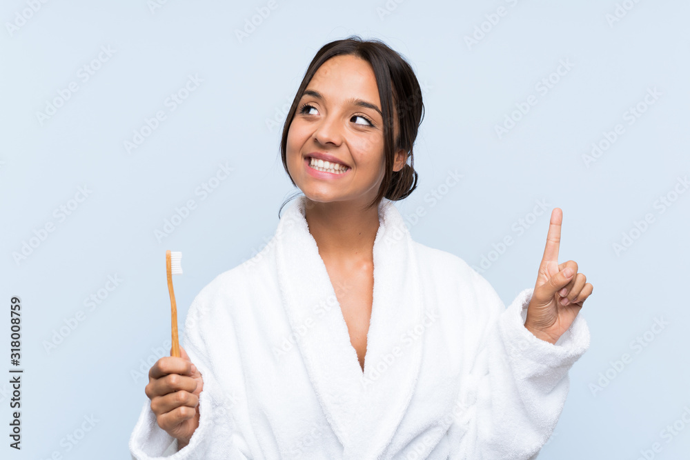 Young brunette woman in bathrobe brushing her teeth over isolated blue background intending to realizes the solution while lifting a finger up Young brunette woman in bathrobe brushing her teeth over isolated blue background intending to realizes the solution while lifting a finger up