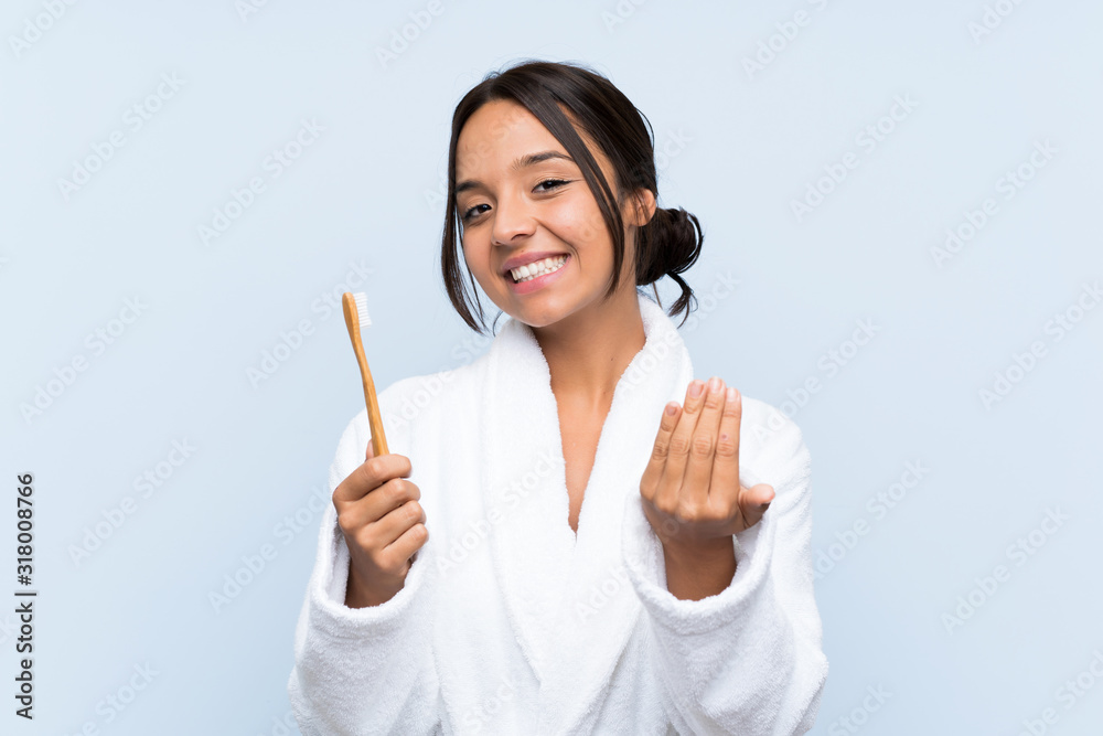 Young brunette woman in bathrobe brushing her teeth over isolated blue background inviting to come with hand. Happy that you came Young brunette woman in bathrobe brushing her teeth over isolated blue background inviting to come with hand. Happy that you came