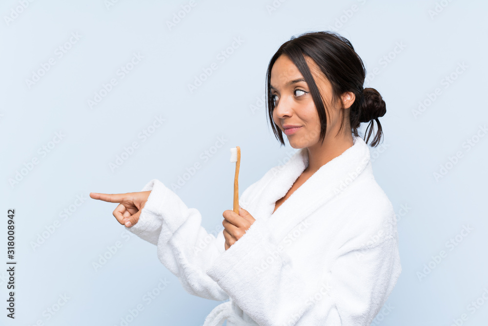 Young brunette woman in bathrobe brushing her teeth over isolated blue background pointing to the side to present a product Young brunette woman in bathrobe brushing her teeth over isolated blue background pointing to the side to present a product