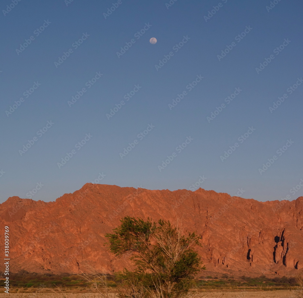 A solitary moon at dusk, contrasting with orange mountains and blue sky