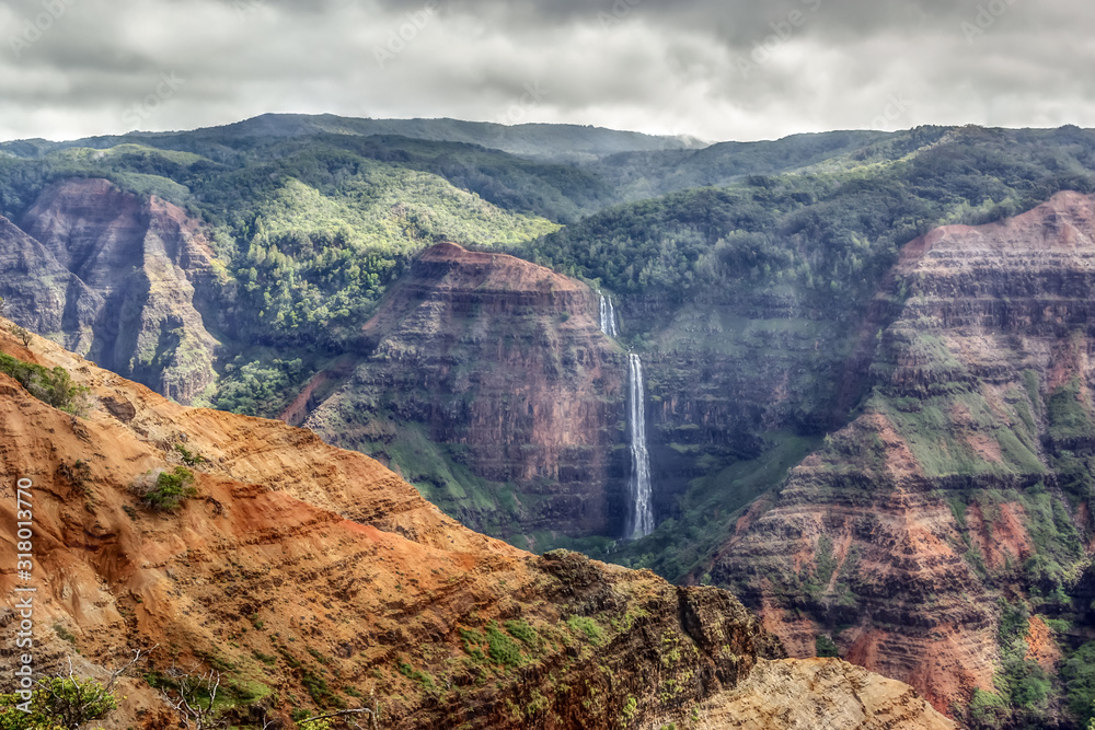 Cloudy grey day at Waimea Canyon State Park, Kauai, Hawaii. The famous Waipo falls plunges to the canyon floor through bands of oxidized basalt rock.