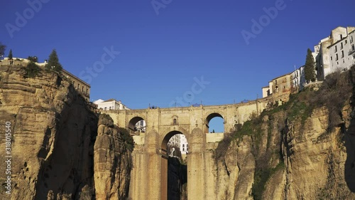 Tilt down shot of stone bridge in Ronda city cliffs and small waterfall under. Water stream under Ronda bridge and cliffs, evening sun