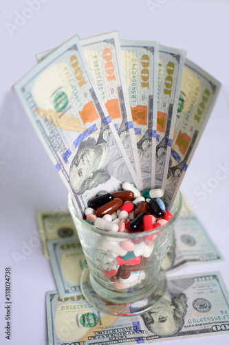 Glass with pills and dollars banknotes. Spending money on pills and expensiveness of medicine concept. Many drug and pill fill in the glass on light background.
