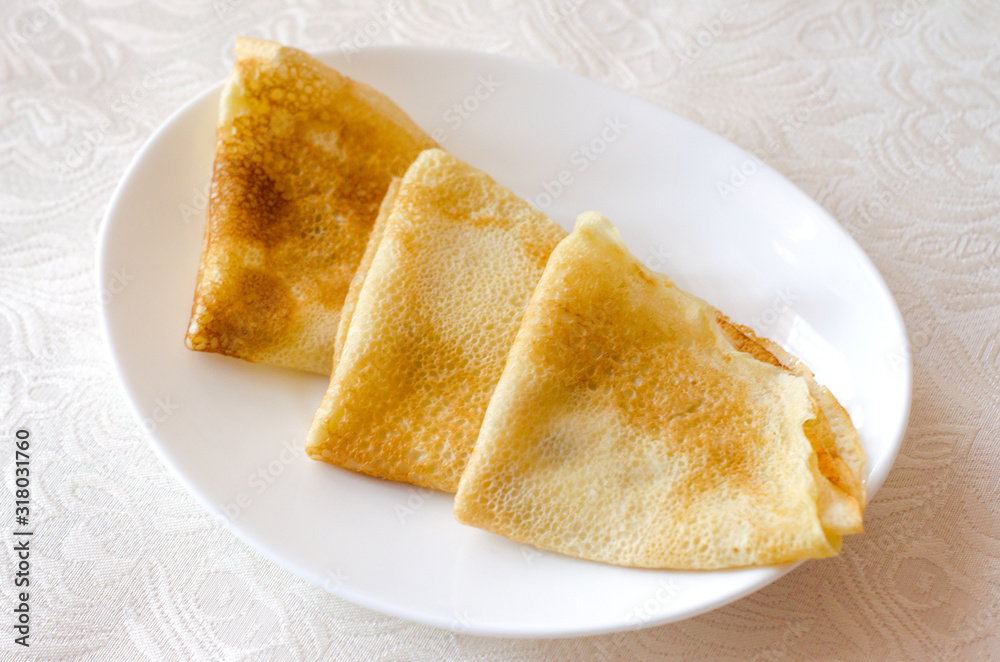 Three pancakes lie on a white plate on a patterned tablecloth. Selective focus. Close up.