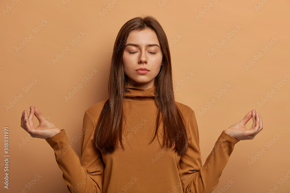 Φωτογραφία Patient relieved woman makes mudra gesture, meditates indoor, tries to relax, breathes deeply and reaches nirvana, closes eyes, wears brown casual jumper