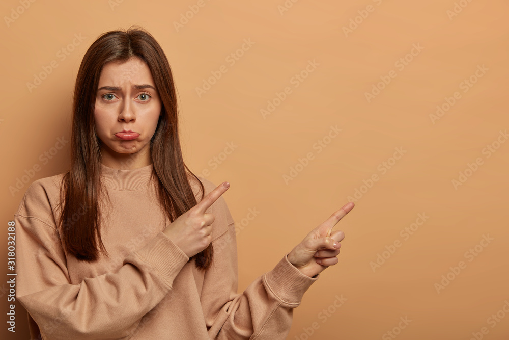 Indoor shot of unhappy disappointed woman points on blank space, missed opportunity, asks your opinion, makes perplexed choice, purses lower lip, isolated over brown background, expresses regret Indoor shot of unhappy disappointed woman points on blank space, missed opportunity, asks your opinion, makes perplexed choice, purses lower lip, isolated over brown background, expresses regret