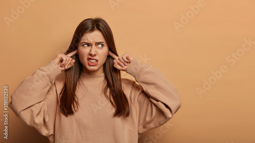 Quadro em tela Horizontal view of young irritated woman uwills hearing bothering noise, plugs ears and frowns face from anger, annoyed by loud music, wears sweatshirt, stands over beige wall