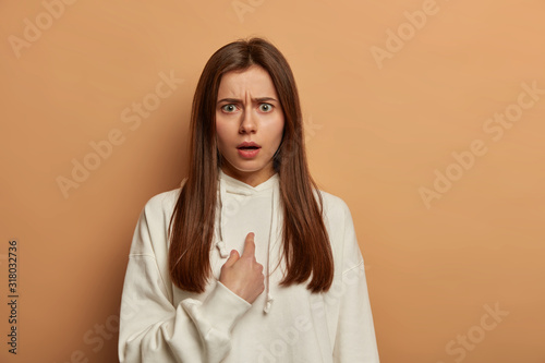 Excuse me. Puzzled indignant woman points at herself, frowns discontentedly face, being confused in doing something wrong, asks question, wears snow white sweatshirt, poses over beige studio wall photo