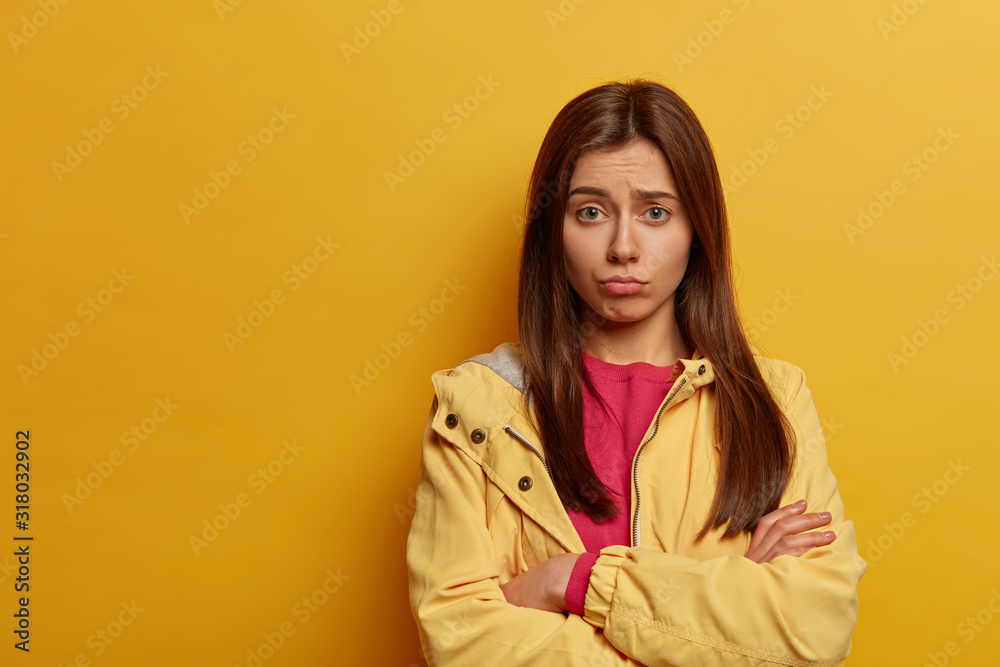 Intense upset woman keeps hands crossed, regrets missing interesting chance, frowns face, looks in dissatisfaction, wears pink jumper and yellow anorak, stands indoor, copy space area on left Intense upset woman keeps hands crossed, regrets missing interesting chance, frowns face, looks in dissatisfaction, wears pink jumper and yellow anorak, stands indoor, copy space area on left