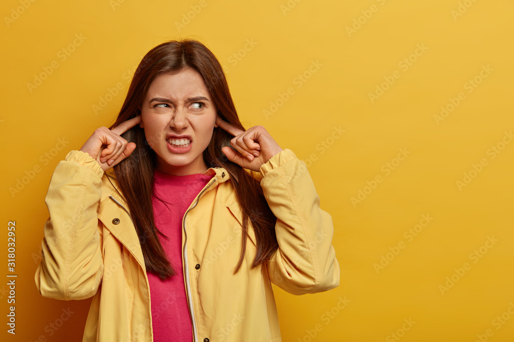 Φωτογραφία Annoyed dark haired young woman plugs fingers into ear holes, clenches teeth, avoids loud annoying noise, wears yellow anorak, stands indoor, hears heartbreaking scream