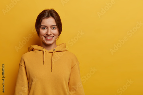 Teenage girl with pleasant smile, has dark combed hair, wears sweatshirt, has healthy skin, stands against yellow background, copy space for your advertisement. Youth, style and emotions concept